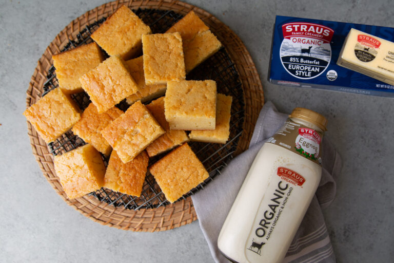 Squares of golden cornbread arranged on a cooling rack and woven tray, photographed alongside a bottle of Straus Family Creamery Organic Eggnog and a package of Straus Organic European Style Butter.