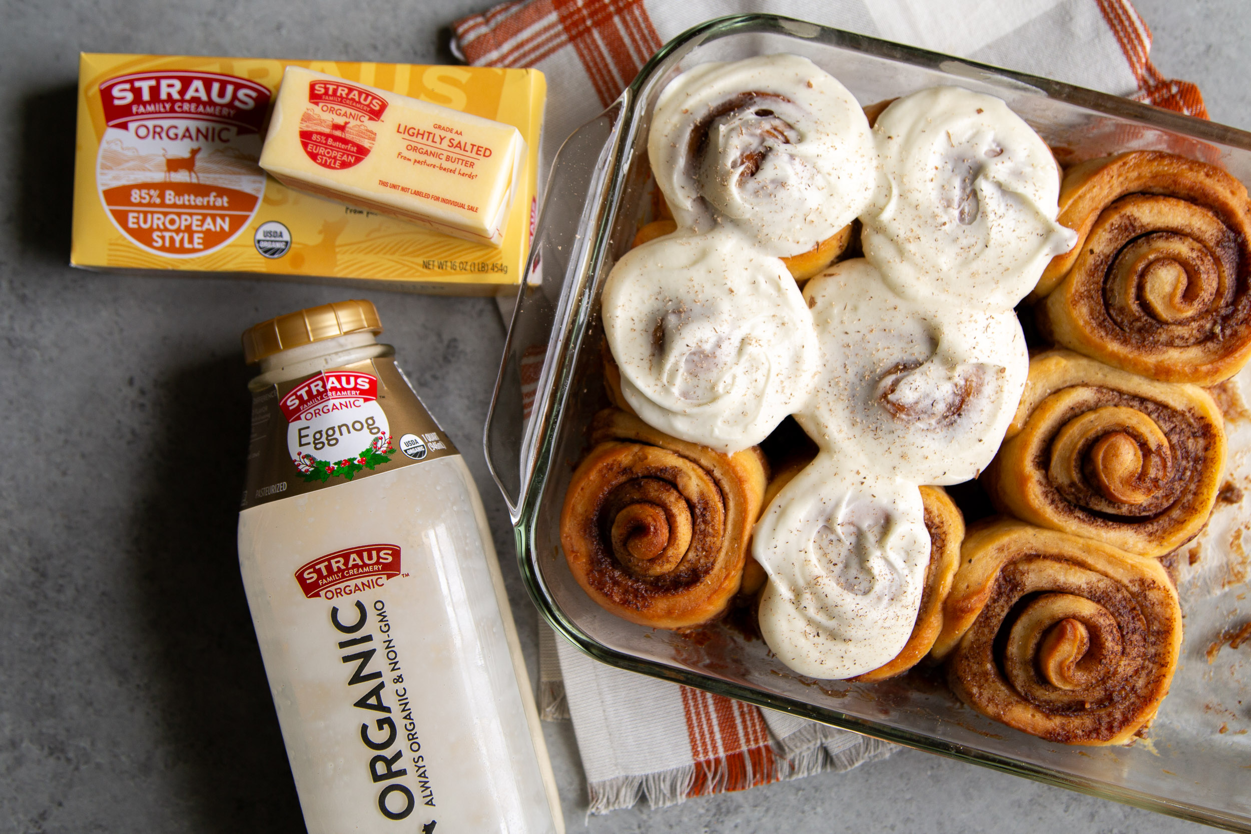 Glass dish of frosted cinnamon rolls next to Straus Family Creamery organic butter and organic eggnog