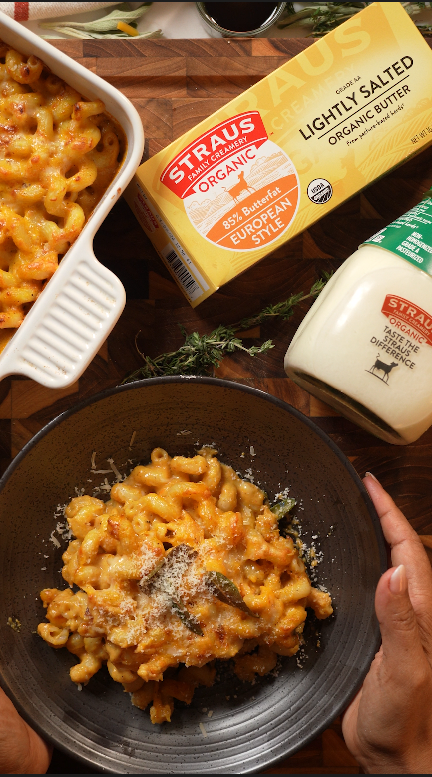 Overhead view of creamy baked macaroni and cheese served in a dark bowl and white baking dish, surrounded by Straus Family Creamery organic lightly salted European-style butter and organic milk on a wooden board.