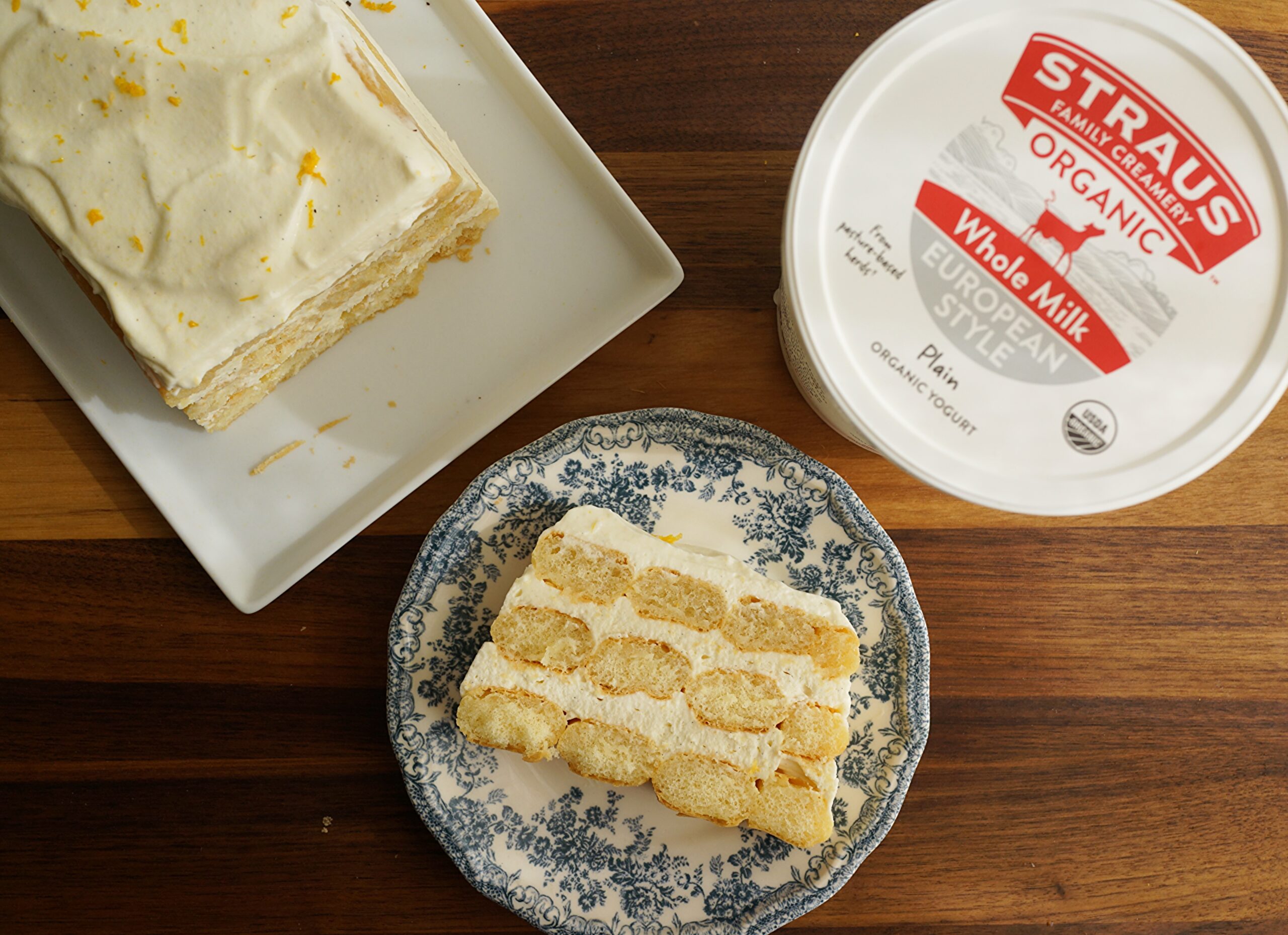 Slice of lemon yogurt tiramisu on a blue patterned plate beside a larger portion and a tub of Straus Organic Whole Milk European Style Plain Yogurt on a wooden table.