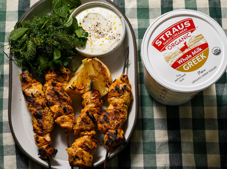 Grilled yogurt-marinated chicken skewers served with fresh herbs, charred lemon halves, and a bowl of yogurt dip beside a container of Straus Organic Whole Milk Greek Yogurt.