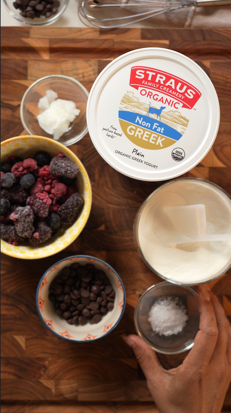 Top-down view of ingredients on a wooden cutting board, including a container of Straus Organic Nonfat Greek Yogurt, frozen mixed berries, chocolate chips, coconut oil, gelatin powder, salt, and a whisk.