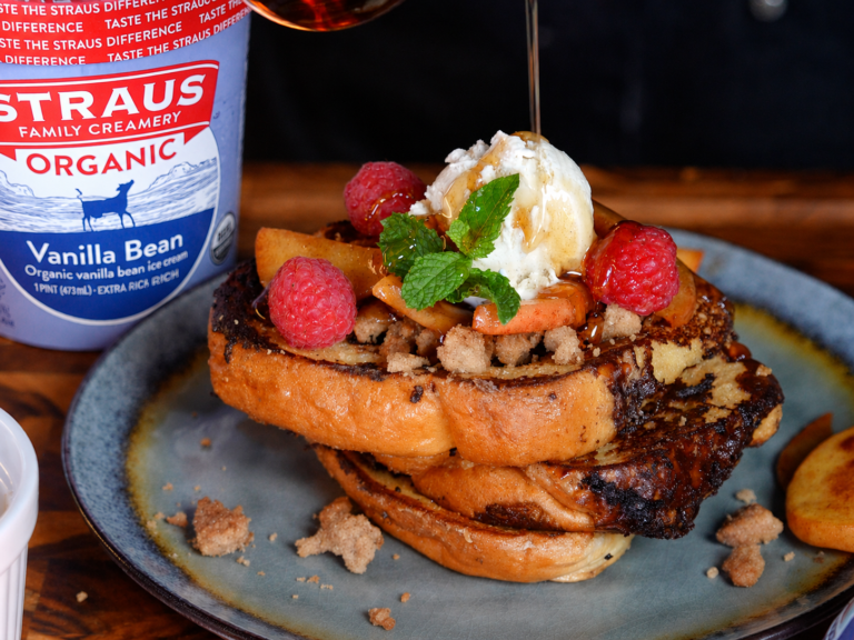 Peaches & cream French toast topped with caramelized peaches, fresh raspberries, mint, buttery crumble, and a scoop of vanilla bean ice cream, with caramel sauce being drizzled over the top, served on a blue plate next to a container of Straus Family Creamery Organic Vanilla Bean Ice Cream.